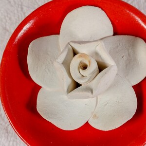 May include: A white ceramic flower shaped diffuser sits on a red ceramic dish. The flower has five petals and a center that resembles a rose.