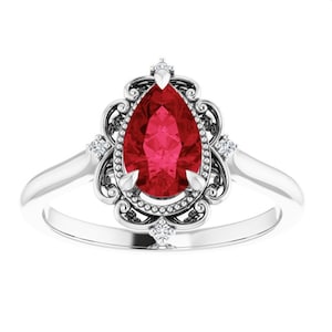 May include: A silver ring featuring a pear-shaped, vibrant red gemstone. The gemstone is surrounded by a decorative filigree setting with small accent diamonds. The ring has a classic, elegant design.