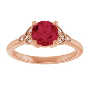 May include: Rose gold ring featuring a large, round, ruby-red gemstone. The ring has a Celtic knot design on each side of the center stone, accented with small, clear gemstones. A classic and elegant piece of jewelry.