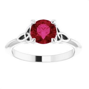 May include: A silver ring featuring a large, round, deep red gemstone. The ring has a Celtic knot design on each side of the gemstone, adding a unique touch. The band is smooth and polished, creating a classic and elegant look.
