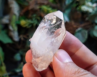 May include: A clear quartz crystal with a double-terminated point, also known as a double-ended crystal. The crystal is held in a hand, showcasing its clarity and facets.