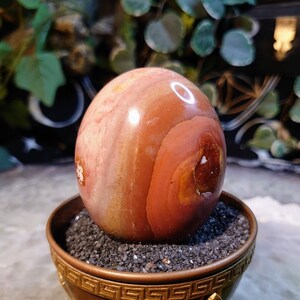 May include: A polished, red and white agate sphere sits on a copper stand with a Greek key design. The sphere is resting on a bed of black sand.