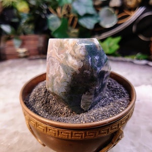 Könnte beinhalten: Eine grüne, weiße und braune Achat-Kristallspitze wird in einem kleinen, goldfarbenen, dekorativen Topf mit einem griechischen Schlüsselmuster präsentiert. Der Topf ist mit dunkelgrauem Sand gefüllt.