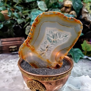 May include: A large, translucent agate slice with brown and white banding is displayed in a copper-coloured stand with a decorative tree design. The agate is lit from below, highlighting its intricate patterns.
