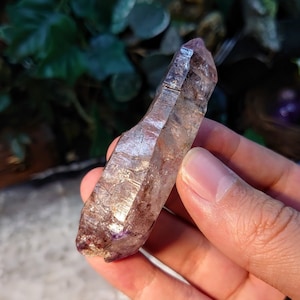 Puede incluir: Una punta de cristal de cuarzo transparente con inclusiones moradas y rosadas. El cristal mide aproximadamente 5 cm de largo y tiene una punta puntiaguda.