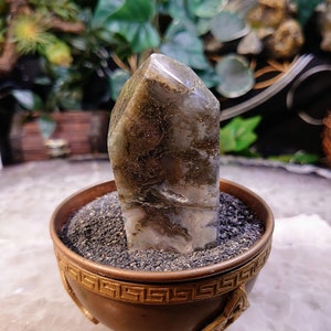 Könnte beinhalten: Eine klare, graue und braune Kristallspitze mit einem metallischen Schimmer, die in einem kleinen, verzierten, kupferfarbenen Topf mit einem geometrischen Muster sitzt.