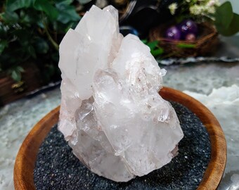 May include: A large, clear quartz crystal cluster with multiple points, resting on a bed of black sand in a wooden bowl.
