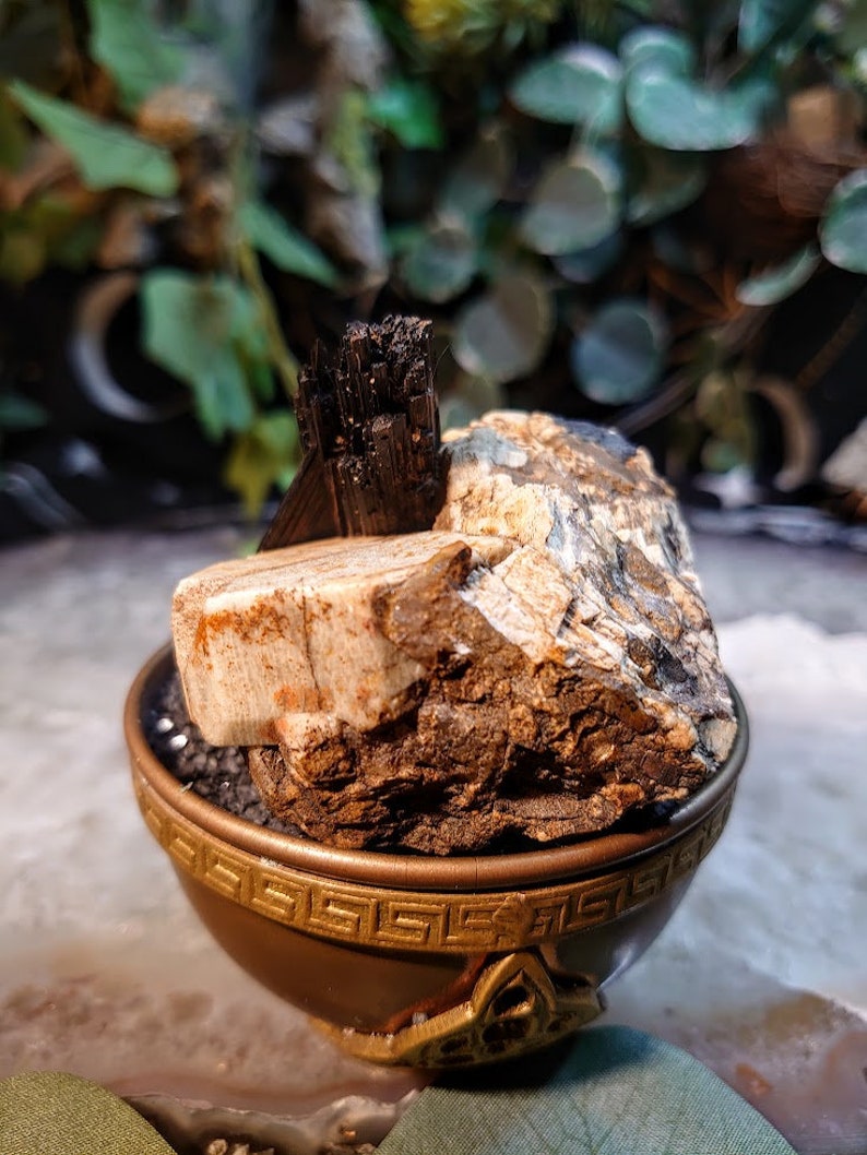 May include: A small, dark brown bowl with a gold-coloured trim and a Greek key design sits on a white surface. Inside the bowl is a large, rough, light brown rock with black and white streaks. A small, black, pointed crystal sits on top of the rock.