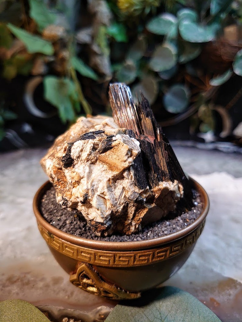May include: A black and brown crystal cluster in a gold-coloured decorative bowl. The crystal cluster is surrounded by black gravel.