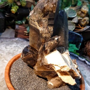 May include: A large, smoky quartz crystal cluster with multiple points, resting on a bed of black sand in a wooden bowl. The crystal has a natural, raw appearance with a brown and clear color.