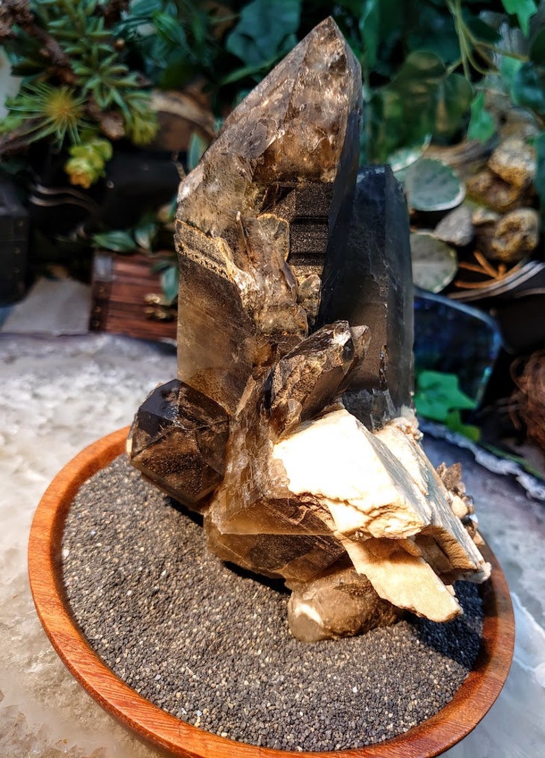 May include: A large, smoky quartz crystal cluster with multiple points, resting on a bed of black sand in a wooden bowl. The crystal has a natural, raw appearance with a brown and clear color.