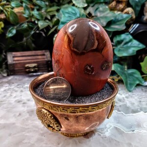May include: A polished red jasper sphere resting in a copper bowl with a gold trim. A US penny is placed in the bowl for scale.