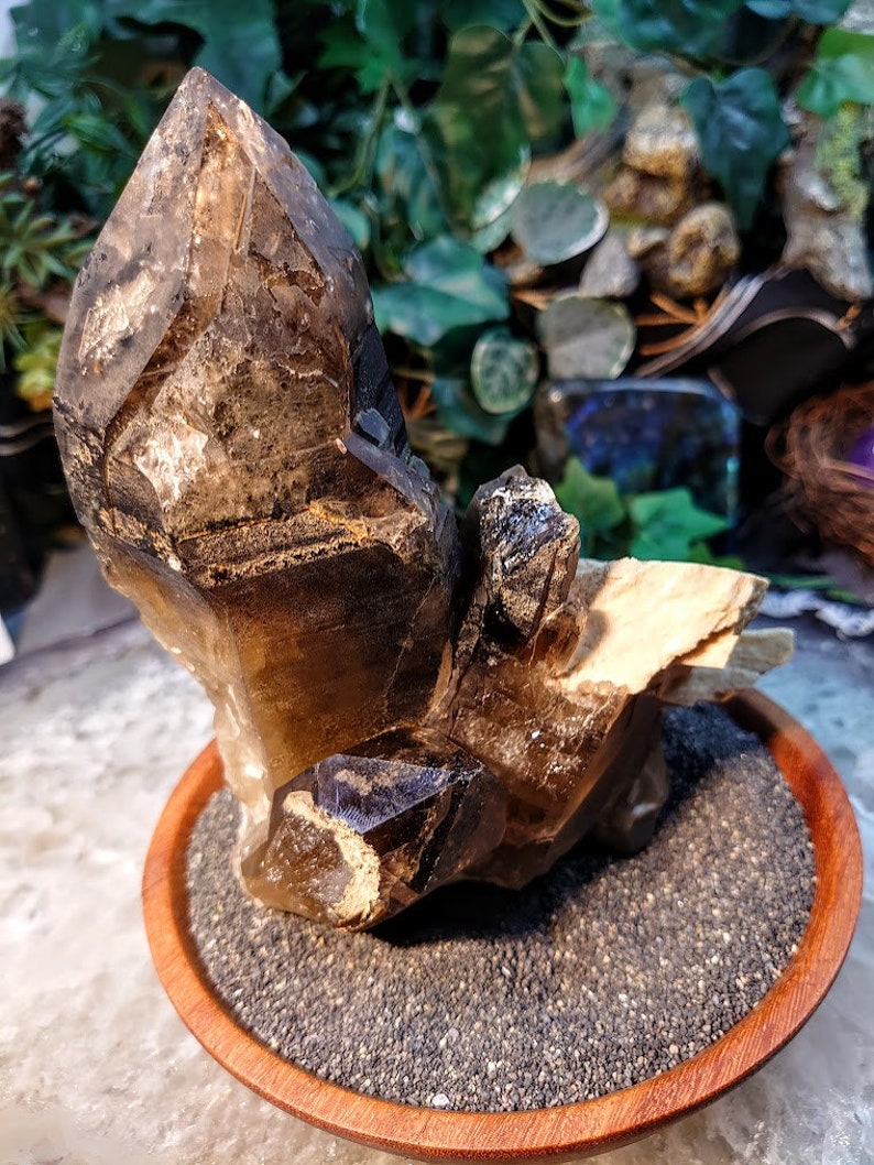 May include: A large, smoky quartz crystal cluster with multiple points, resting on a wooden base filled with black sand. The crystal is translucent and has a brown hue.