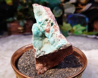 May include: A teal blue and brown crystal specimen on a copper stand with a decorative border. The crystal is sitting on a bed of black sand.