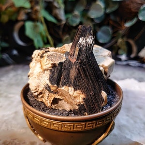 May include: A black and white crystal cluster in a gold-coloured bowl with a Greek key design. The crystal cluster is sitting on a bed of black sand.