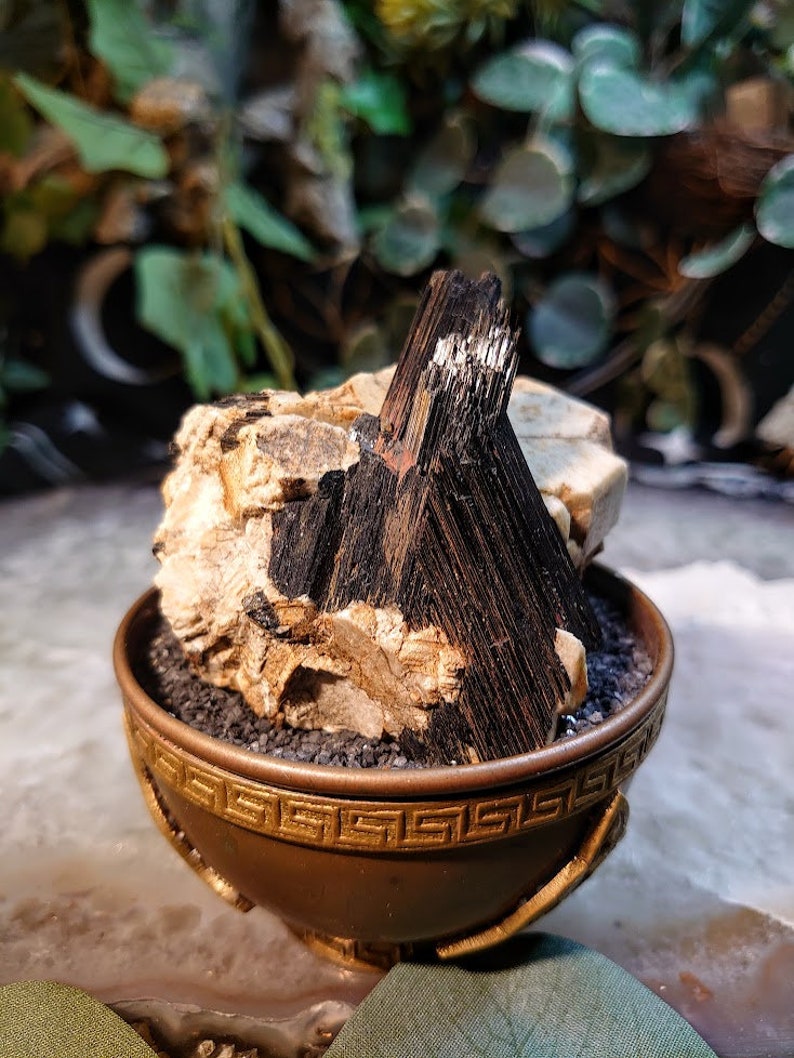May include: A black and white crystal cluster in a gold-coloured bowl with a Greek key design. The crystal cluster is sitting on a bed of black sand.