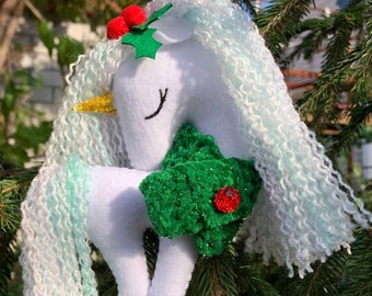 May include: White felt unicorn ornament with a green felt holly leaf and red berries. The unicorn has a long, white and light blue yarn mane and tail.