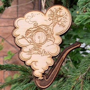May include: A wooden ornament shaped like a pipe with a cloud of smoke coming out of it. The cloud has a detailed image of a hobbit hole with a round door and a tree growing out of the roof.