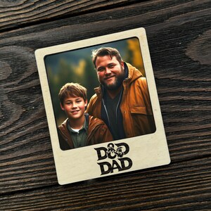 Può includere: Una cornice in legno con una foto di un uomo e un ragazzo, su uno sfondo esterno sfocato. La cornice presenta le parole "D&D DAD" in un carattere stilizzato. La cornice è posizionata su una superficie di legno scuro.