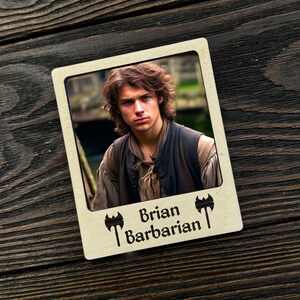 Può includere: Una cornice per foto in legno con una foto di un uomo con i capelli castani e la barba. La cornice ha uno sfondo bianco con il testo "Brian Barbarian" e due simboli di ascia.