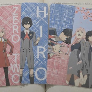 May include: Four anime-themed bookmarks featuring characters from the series. The bookmarks display images of the characters Zero Two and Hiro, with text that reads "ZERO TWO" and "HIRO". The bookmarks are on top of an open book.