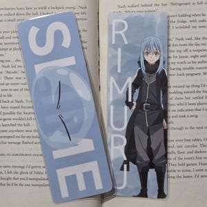 May include: Two bookmarks rest on an open book. One bookmark is light blue with the word "SLIME" in white. The other bookmark features an anime character with the name "RIMURU" in white text.