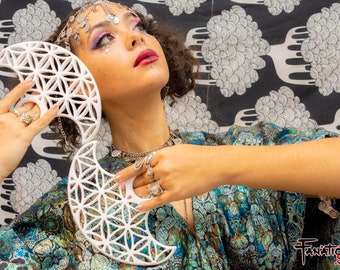 May include: A white ceramic crescent moon with a geometric pattern, held by a person wearing a silver headpiece and a blue and green patterned robe.