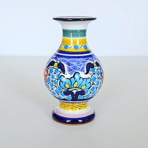 May include: A ceramic vase featuring a hand-painted design with a white base, vibrant blue floral motifs, yellow bands, and green accents. The piece has a flared rim, a rounded body, and a pedestal base with intricate geometric patterns throughout.