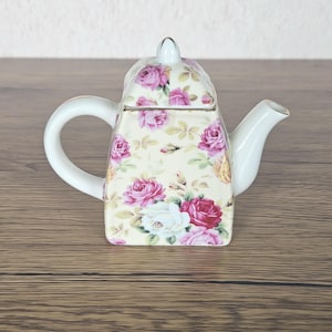 May include: A square-shaped porcelain teapot featuring a cream-colored background decorated with a pattern of pink and white roses and green leaves. The teapot includes a matching lid with a small knob, a curved handle, and a spout.