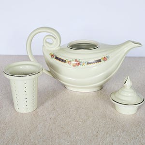 May include: Cream-colored ceramic teapot with a curved handle and spout, a separate tea infuser, and a lid. The teapot features a floral and ribbon design. The infuser and lid have black trim.