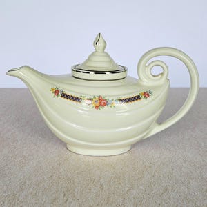 May include: Cream-colored ceramic teapot with a curved handle and spout. The lid is topped with a gold finial. A decorative band of flowers and a navy blue crisscross pattern adorns the body of the teapot. Silver trim accents the lid and spout.