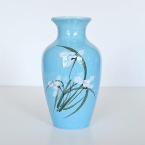 May include: A light blue ceramic vase with a flared rim and a tapered neck. The body of the vase features a hand-painted botanical design consisting of white flower blossoms and long, thin green leaves rendered in a fluid, brushstroke style.