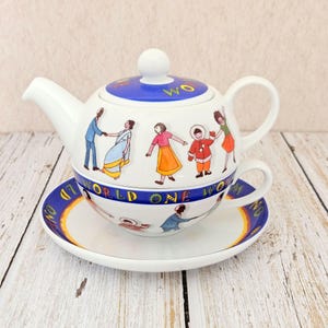 May include: A white ceramic teapot, teacup, and saucer set. The teapot and teacup are decorated with colorful illustrations of people and the words "ONE WORLD WOM". The set is on a white and light brown wooden surface.