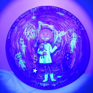 May include: A round, hand-painted artwork with a surreal, glowing design. The artwork features a central figure holding a doll, surrounded by other characters and the text "WHEN YOU'RE SCARED BUT YOU STILL DO IT ANYWAY THAT'S BRAVE."