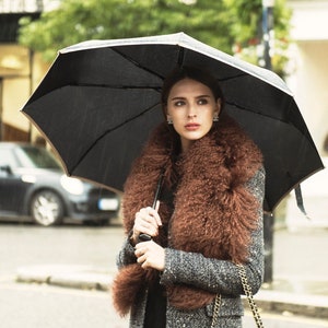 May include: A person shelters from the rain under a black umbrella. They are wearing a grey tweed coat, a brown fur collar, and carrying a black quilted handbag with a gold chain. The background shows a street scene.