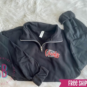 May include: Black quarter-zip sweatshirt with a silver zipper and the word "Chiefs" in red and white lettering. The sweatshirt is laid on a white, fluffy surface. The sleeves are cuffed.