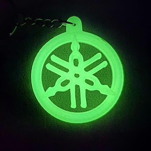 May include: A glowing green Yamaha logo keychain. The keychain features the Yamaha logo within a circular frame, with a chain attached for easy carrying. The design glows brightly against a dark background.
