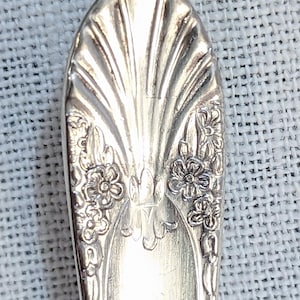 May include: Close-up of a silver spoon with a decorative floral and fan design. The spoon's bowl features a central fan-like pattern with floral accents on either side. The spoon is made of a shiny, reflective metal.