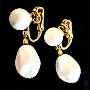 May include: A pair of clip-on earrings with a gold-tone metal design. Each earring features a white, round bead at the top and a larger, teardrop-shaped white bead hanging below. The earrings are set against a black background.
