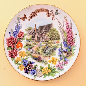 May include: A white porcelain plate with a gold rim features a colorful illustration of a cottage garden scene. The plate is decorated with flowers, birds, and a rabbit. The text "ENGLAND" is written in gold lettering on the plate.