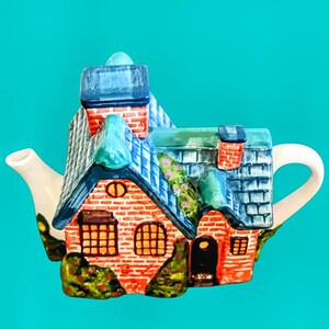 May include: A whimsical ceramic teapot shaped like a house. The teapot features a red brick facade, blue roof, and a white handle and spout. The windows are a warm yellow, and the chimney is red. The overall design is detailed and colorful.
