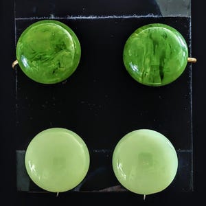 May include: Four round earrings in shades of green are displayed on a black surface. Two earrings are a vibrant, marbled green, while the other two are a lighter, solid green. The earrings have gold-colored posts.