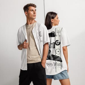 May include: Two people wearing white baseball jerseys and t-shirts. The jersey has the text "THE" and "ANG" with a circular design. The t-shirt has a black and white graphic design with the text "Jackson Wang".