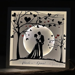 Könnte beinhalten: Eine weiße Holz-Schattenbox mit einer Silhouette eines Paares, das unter einem Mond mit roten Herzen steht, die um sie herum verstreut sind. Die Box wird von hinten beleuchtet, wodurch ein leuchtender Effekt entsteht. Der Text "Nicole & Daniel 21.09.2024" ist unten eingraviert.
