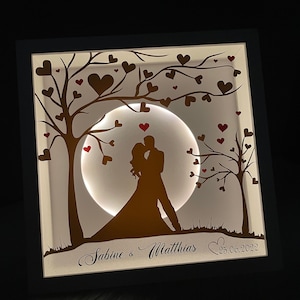 Könnte beinhalten: Ein weißer Holzrahmen mit einem ausgeschnittenen Design von zwei Silhouetten von Menschen, die sich unter einem Mond mit einem Baum auf jeder Seite küssen. Der Mond wird mit einem warmweißen Licht beleuchtet. Der Rahmen trägt den Text "Sabine & Matthias 25.06.2022" unten.