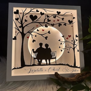 Könnte beinhalten: Beleuchtetes Kunstwerk in einem Rahmen, das ein älteres Paar zeigt, das auf einer Bank unter Bäumen mit herzförmigen Blättern sitzt. Die Namen "Lieselotte & Horst" und "50 Jahre" sind enthalten.