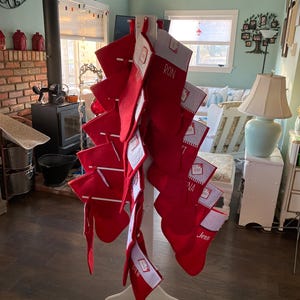 Puede incluir: Un soporte de calcetines de madera blanco con calcetines rojos y blancos colgando de él. Los calcetines están personalizados con nombres como "Ron", "Jenna" y "Olivia".