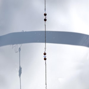 Può includere: Un ornamento verticale sospeso caratterizzato da una catena in metallo scuro intervallata da perline rotonde marroni e perline più piccole trasparenti e iridescenti. La catena termina con una goccia di cristallo trasparente sfaccettato.