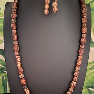 May include: A necklace and earring set featuring brown and tan patterned beads. The necklace is displayed on a black bust, with the earrings hanging beside it. The beads have a unique design, and the earrings have gold-colored hooks.
