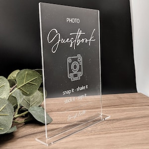 May include: A clear acrylic sign with the text "PHOTO GUESTBOOK" in white. The sign also features a camera icon and the text "snap it - shake it - stick it - sign it".